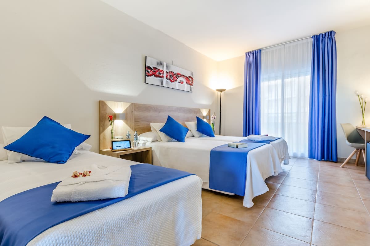 A hotel room with two beds, each with blue bedding and pillows, a bedside table with a lamp, and a large window with blue curtains.