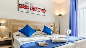 Habitación de hotel con dos camas, cabecera de madera, ropa de cama azul y blanca, un cuadro en la pared y una mesita de noche con una lámpara y una botella.