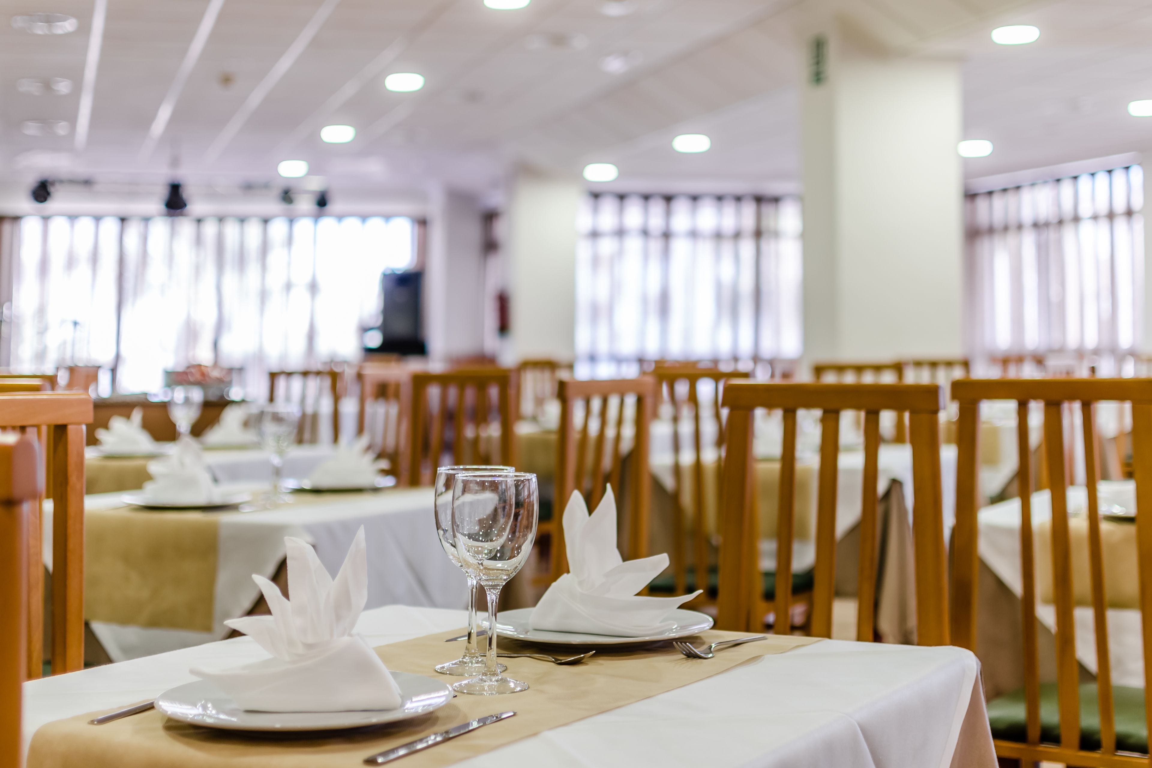 Un comedor con mesas dispuestas para el servicio, con manteles blancos, servilletas dobladas y copas de vino vacías.