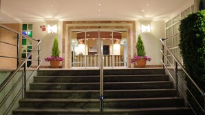 Entrada de hotel con una escalinata de mármol, barandillas metálicas y plantas en macetas.