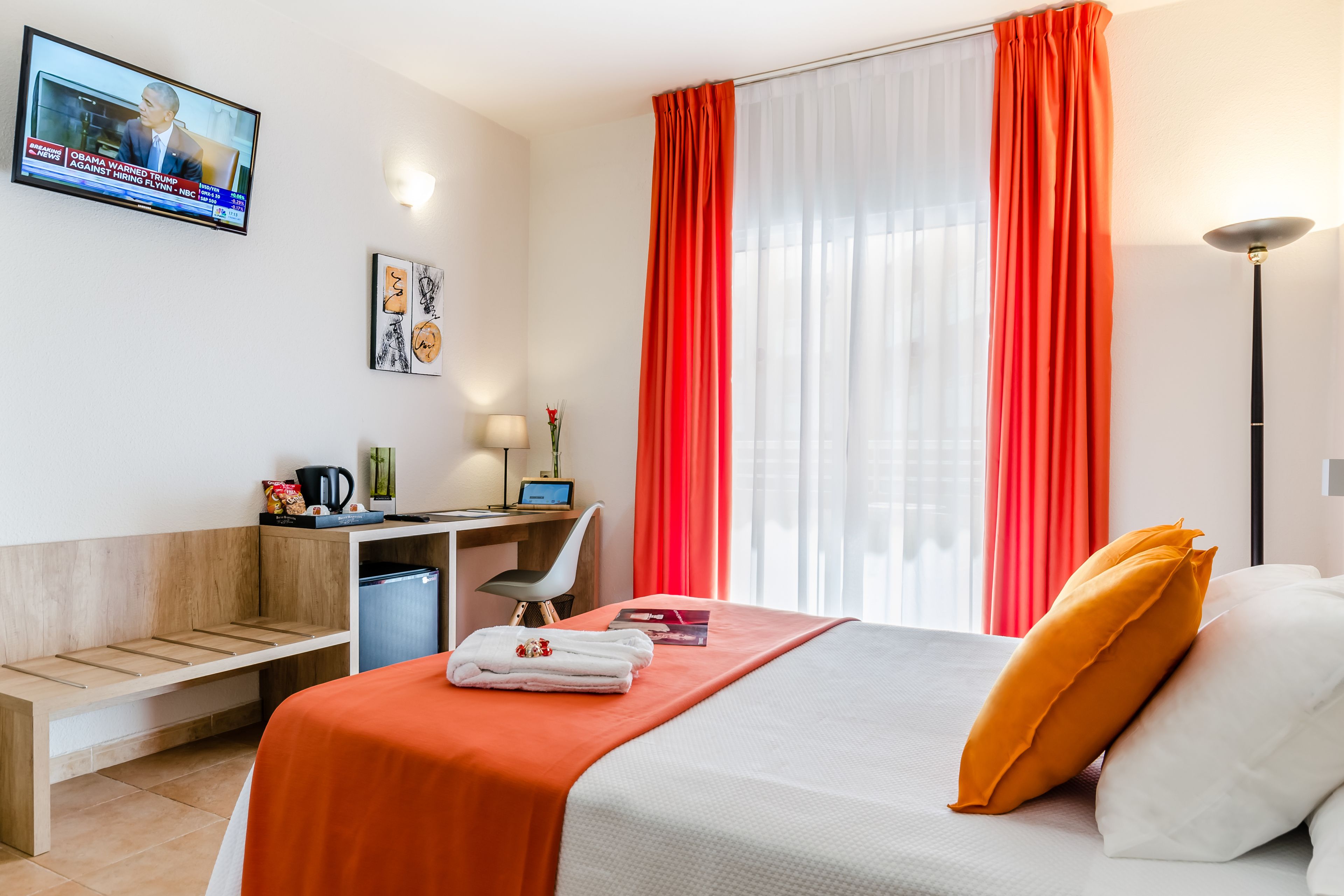 Una habitación de hotel con cama, escritorio, silla, televisión y ventana con cortinas.