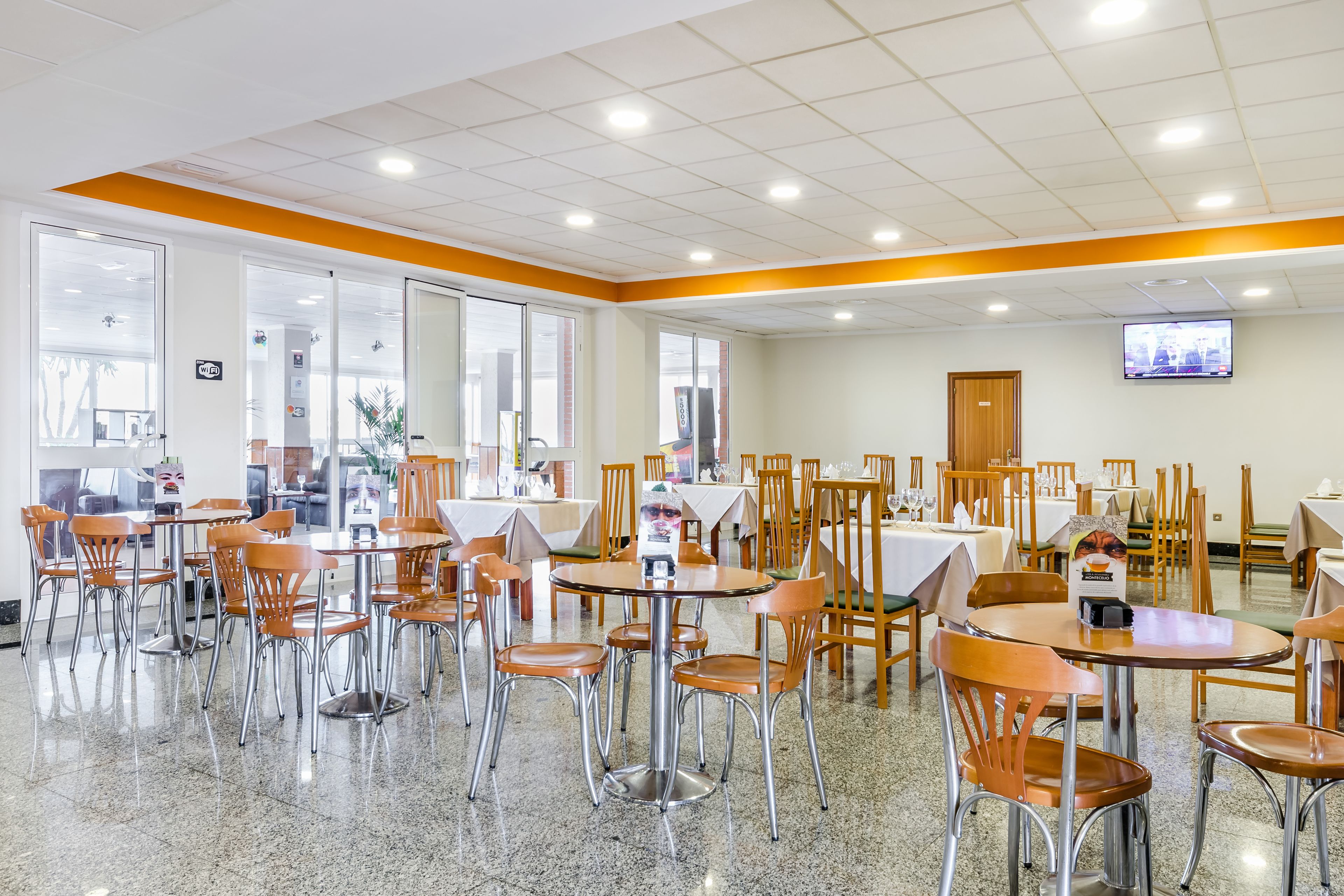 A spacious dining area with multiple tables and chairs, large windows, and a television mounted on the wall.