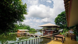 Una terraza de madera con vistas a una masa de agua tranquila, un pequeño edificio con techo y abundante vegetación.