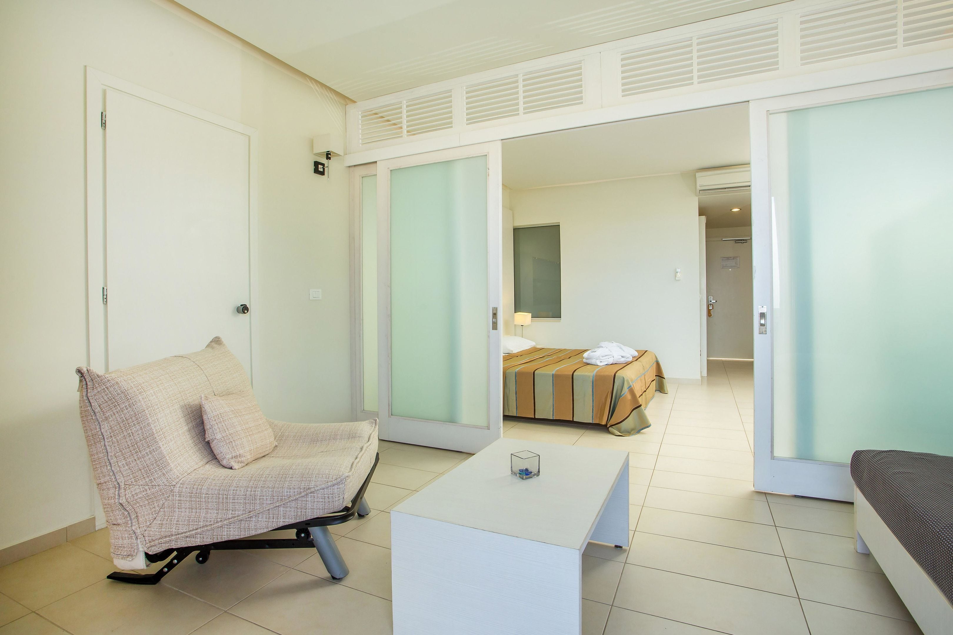 A hotel room with a single bed, a chair, a small table, and a sliding door leading to another room.