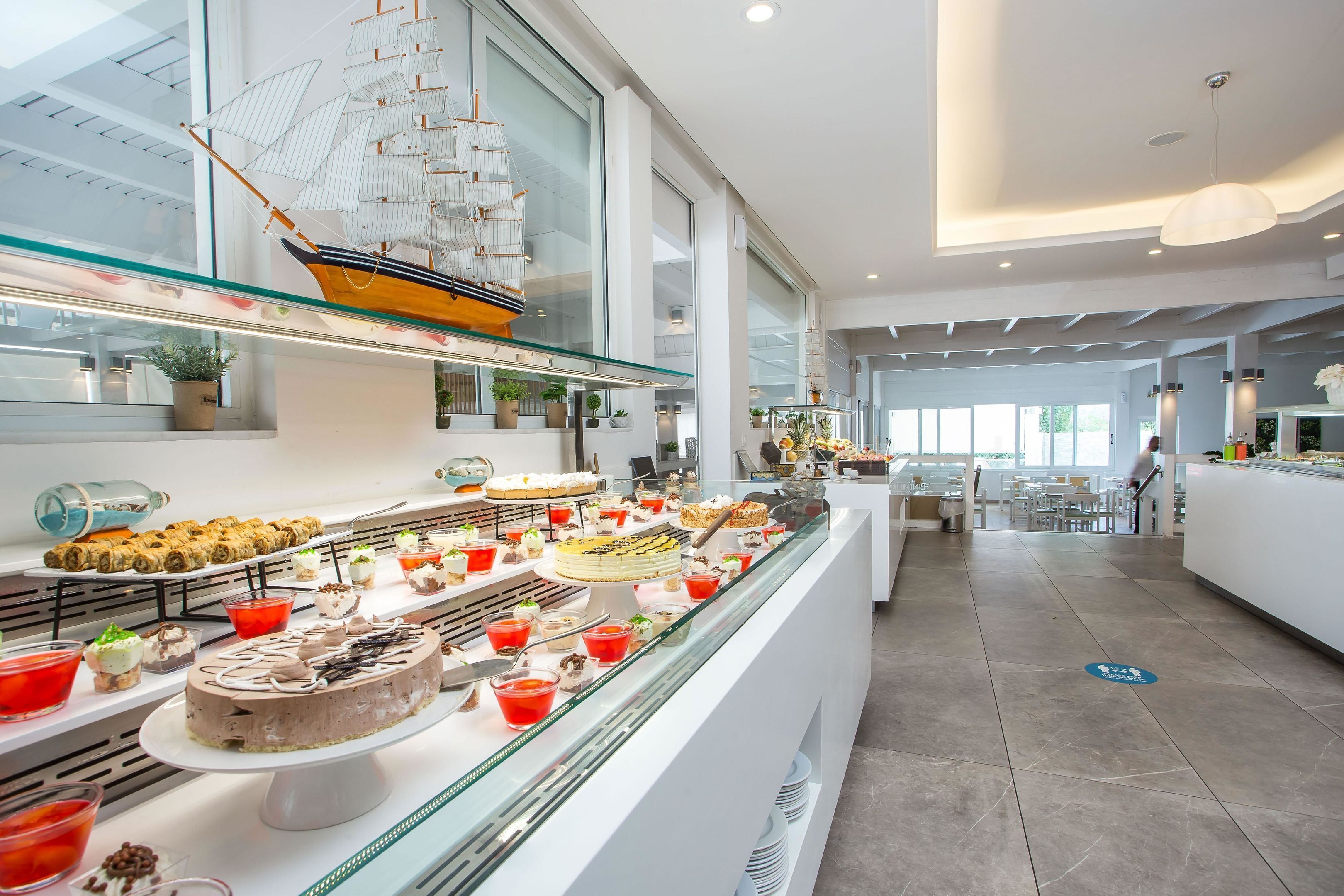 A modern hotel lobby with a glass display case filled with various desserts and pastries, a large ship model, and a seating area with tables and chairs.