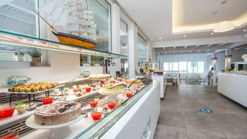 A modern hotel lobby with a glass display case filled with various desserts and pastries, a large ship model, and a seating area with tables and chairs.