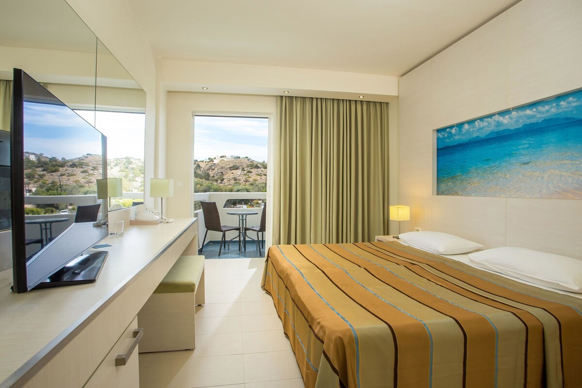 A hotel room with a bed, a desk with a computer, a chair, a window with curtains, and a painting on the wall.