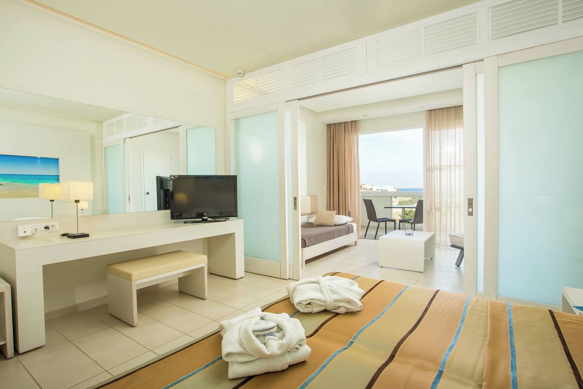 A hotel room with a bed, a desk with a television, a bench, and a view of the outdoors.