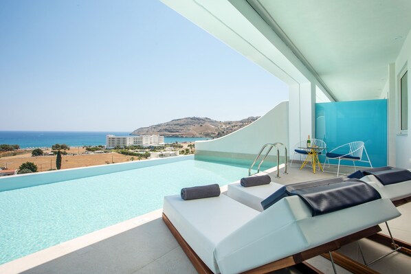 A modern infinity pool with a view of the sea and a hillside. There are lounge chairs with towels and a table with chairs.