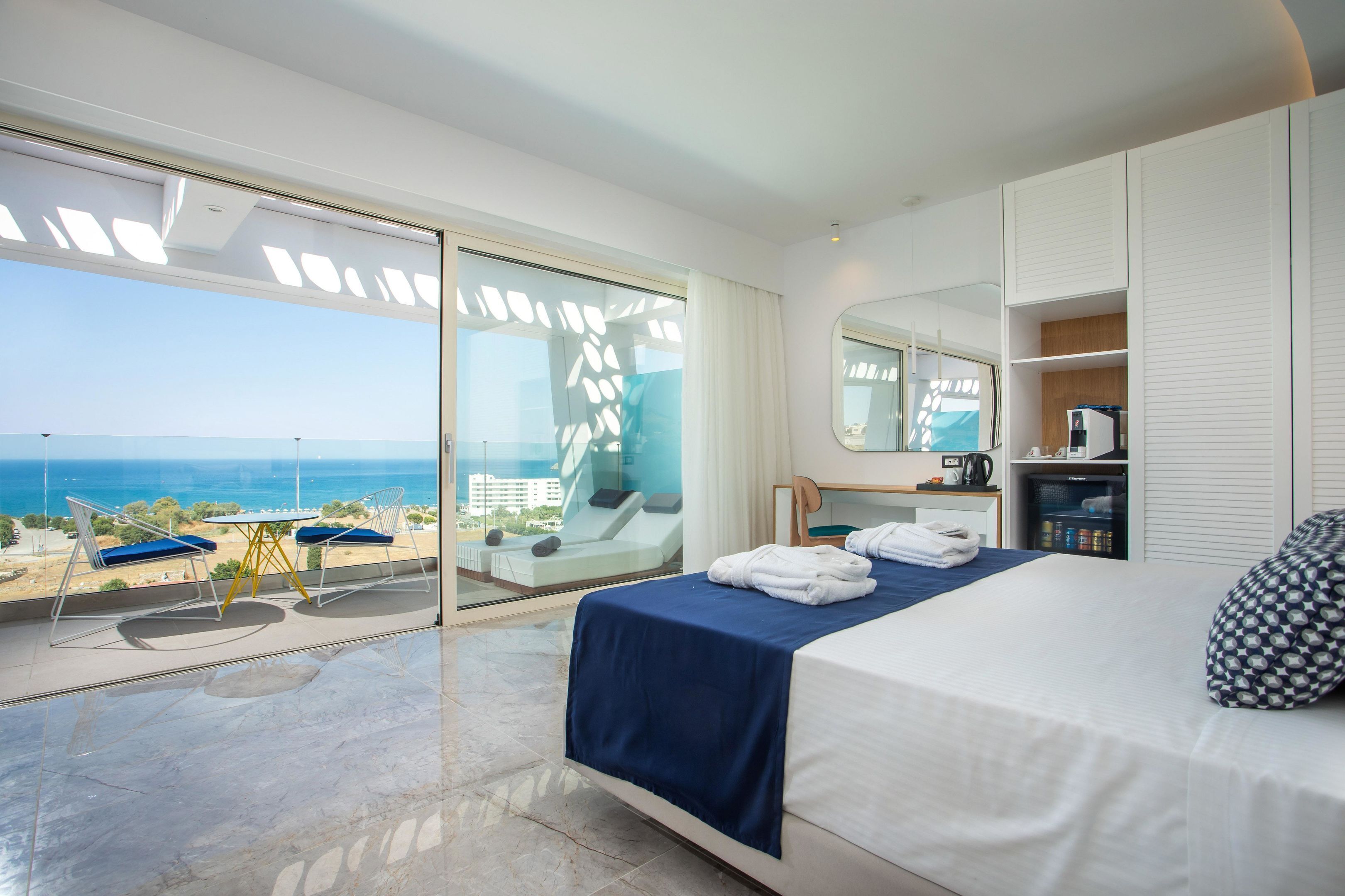 A modern hotel room with a balcony overlooking the sea, a bed with blue bedding, a desk with a chair, and a wardrobe.