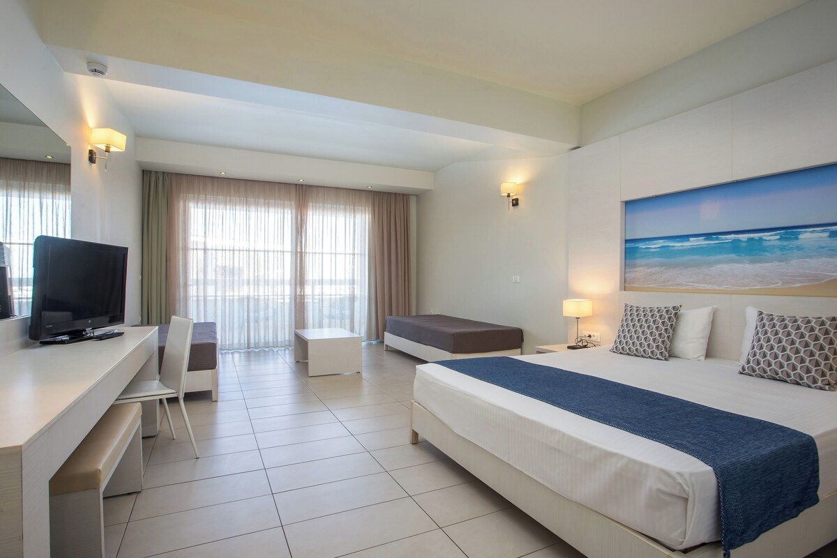 A hotel room with a bed, a desk with a chair, a television, and a large window with curtains.