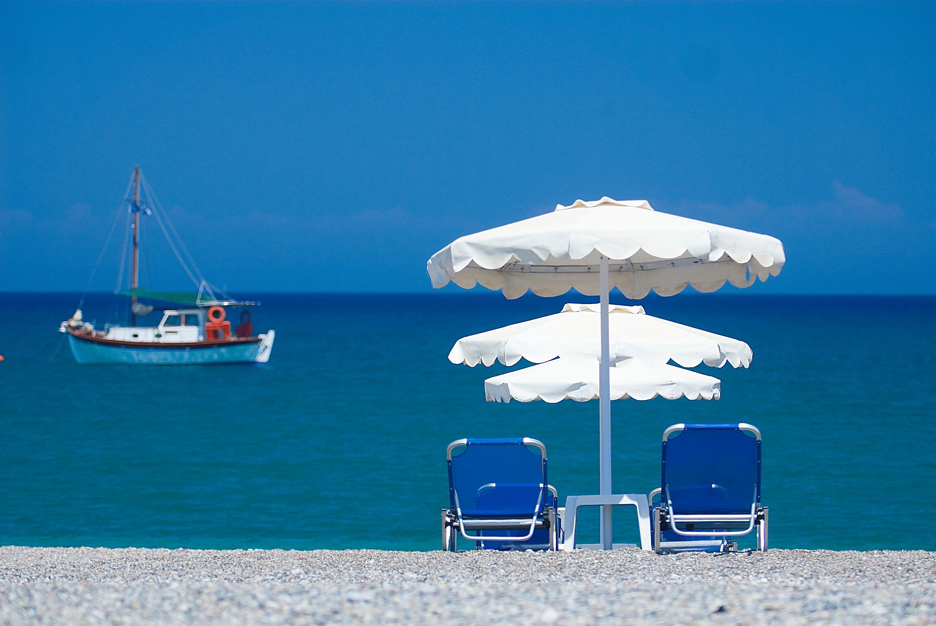A beach with two blue chairs under a white umbrella, a boat in the background, and a clear blue sky.