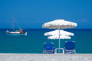 A beach with two blue chairs under a white umbrella, a boat in the background, and a clear blue sky. - Lindos White Hotel & Suites (Rhodes)