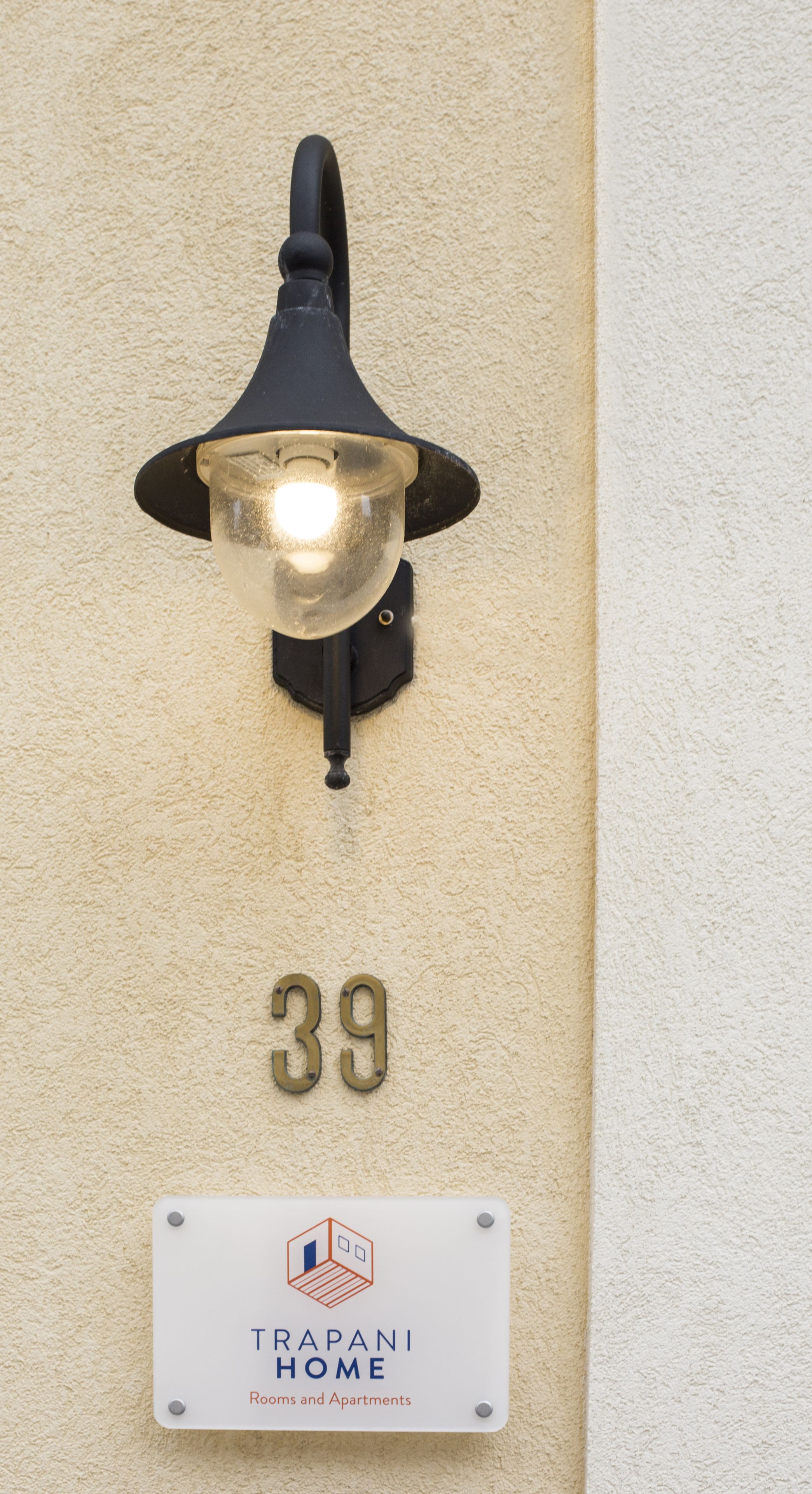 Una lámpara de pared con pantalla de vidrio transparente, una placa con el número '39' y un cartel de 'TRAPAN HOME' que indica habitaciones y apartamentos.