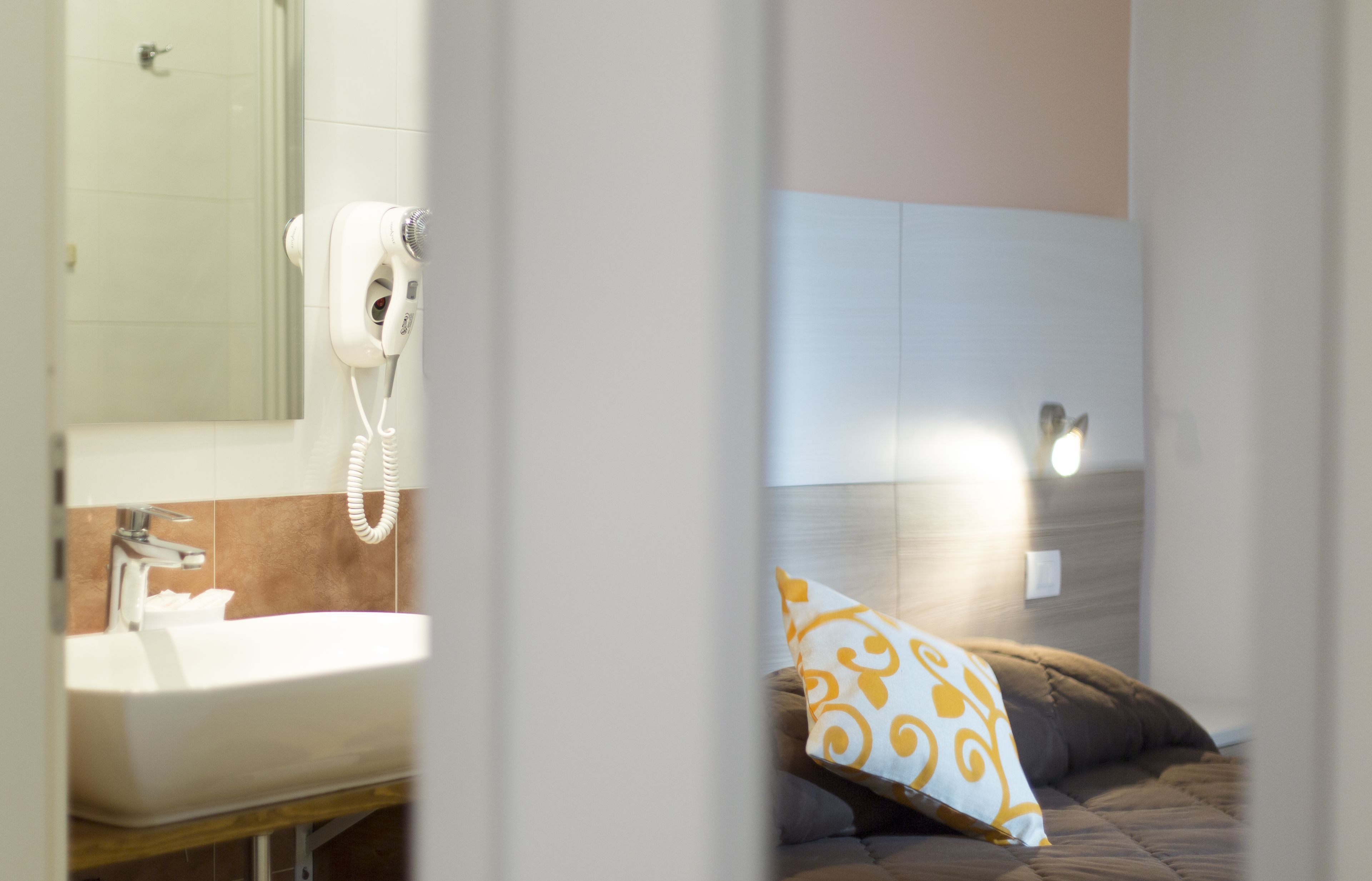 A bathroom with a sink, mirror, and hairdryer. There is a bed with a pillow in the adjacent room.