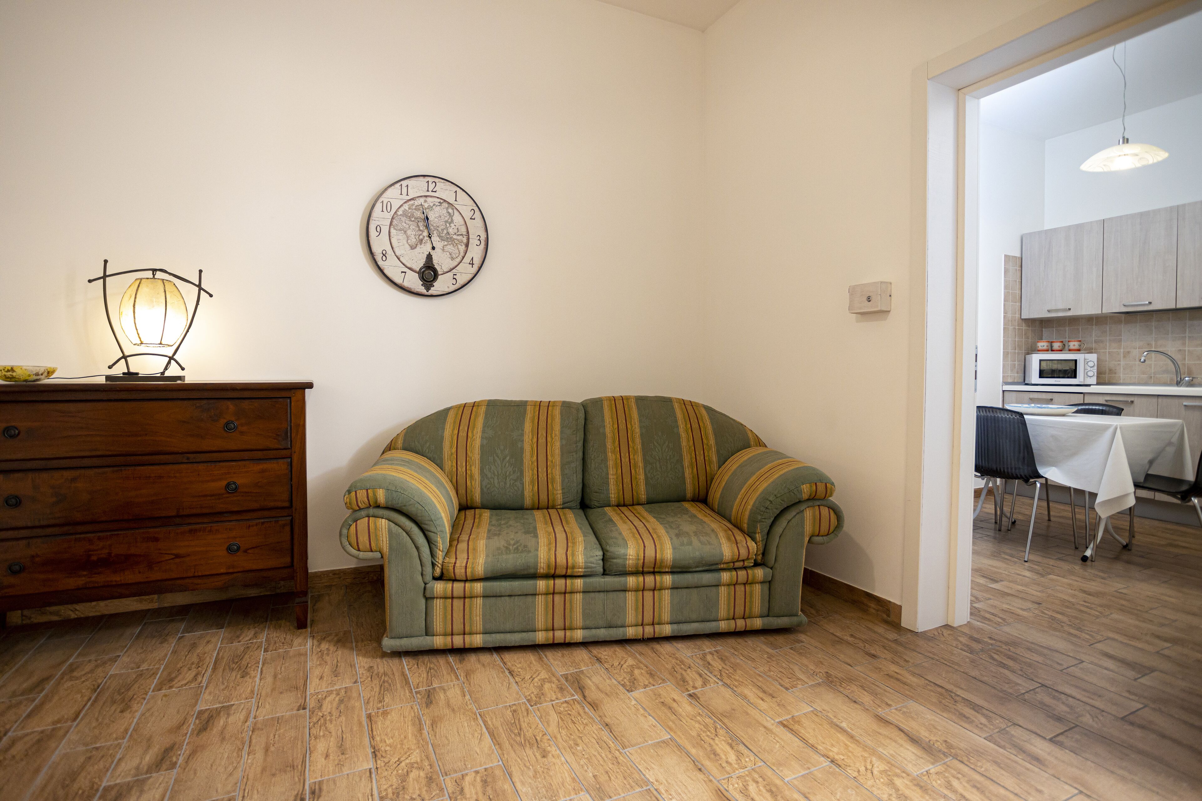 A living room with a sofa, wooden dresser, wall clock, and a kitchen visible through an open doorway.