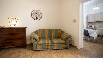 A living room with a sofa, wooden dresser, wall clock, and a kitchen visible through an open doorway.