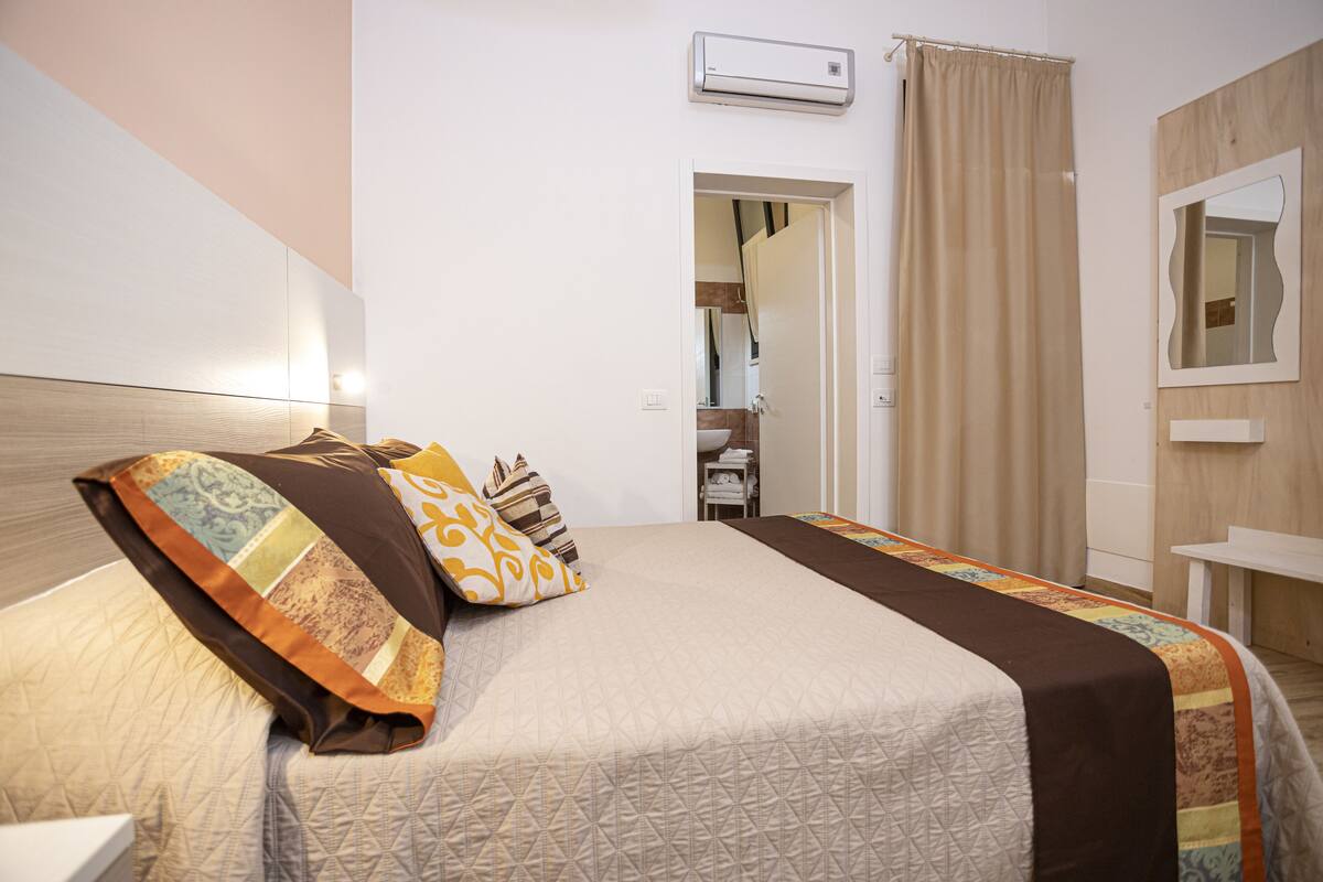 Habitación de hotel con cama, mesita de noche, espejo y ventana con cortinas.