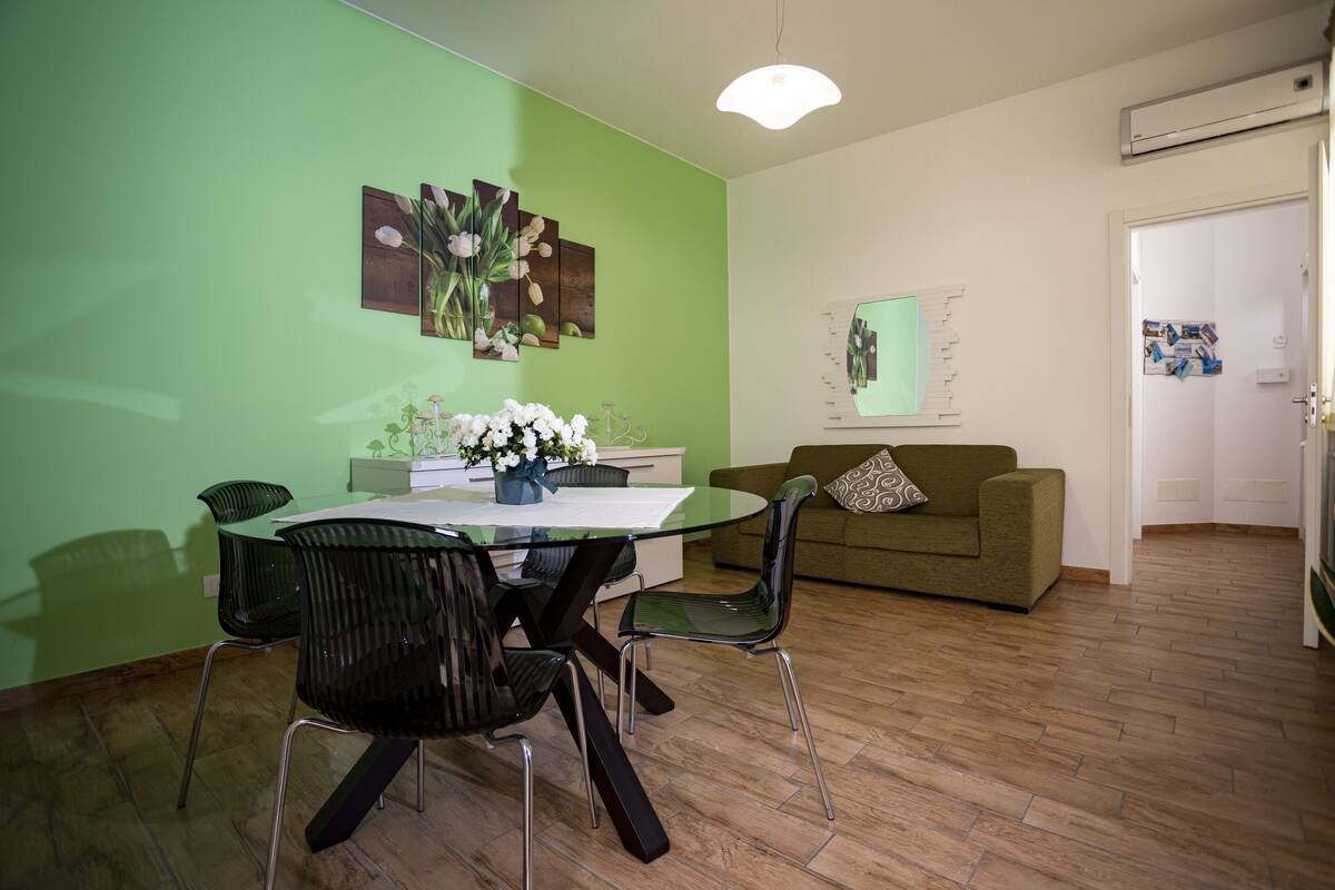 Un área de comedor con una mesa y sillas de vidrio, una pared verde con obras de arte, un sofá y una puerta con un tablón de anuncios.