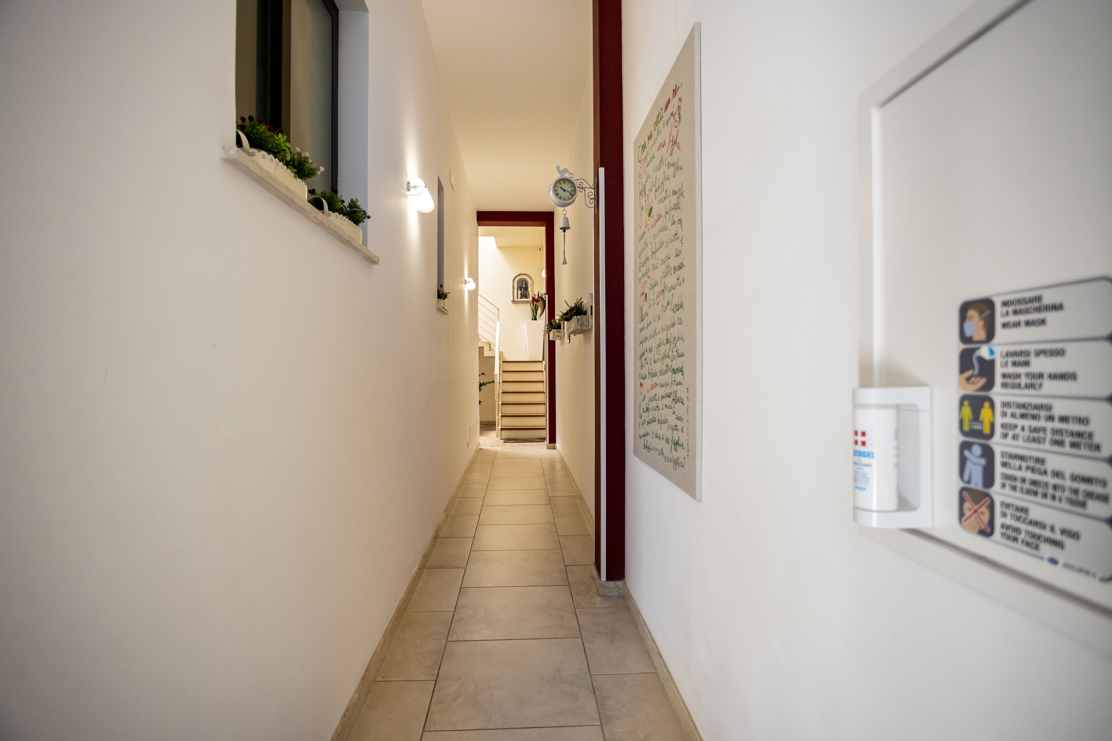 A narrow hallway with a tiled floor, a window with plants, a clock, and a wall with health and safety instructions.