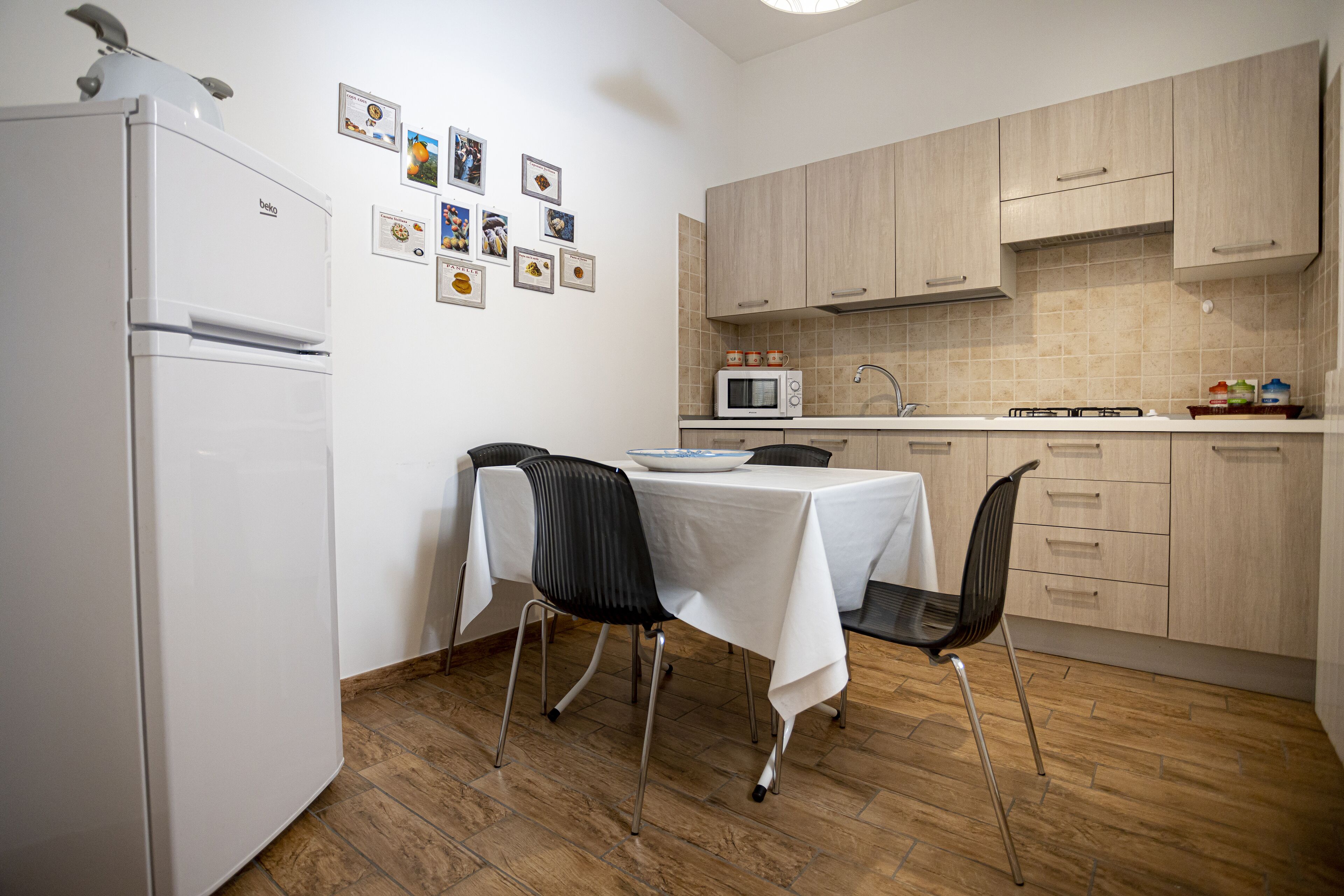 A compact kitchen with a refrigerator, microwave, and wooden cabinets. There is a dining table with chairs and a wall decorated with framed pictures.
