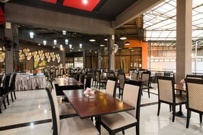 A spacious restaurant with multiple dining tables and chairs, a high ceiling with exposed beams, and a large window allowing natural light. - Prelude Hotel (Kanchanaburi)