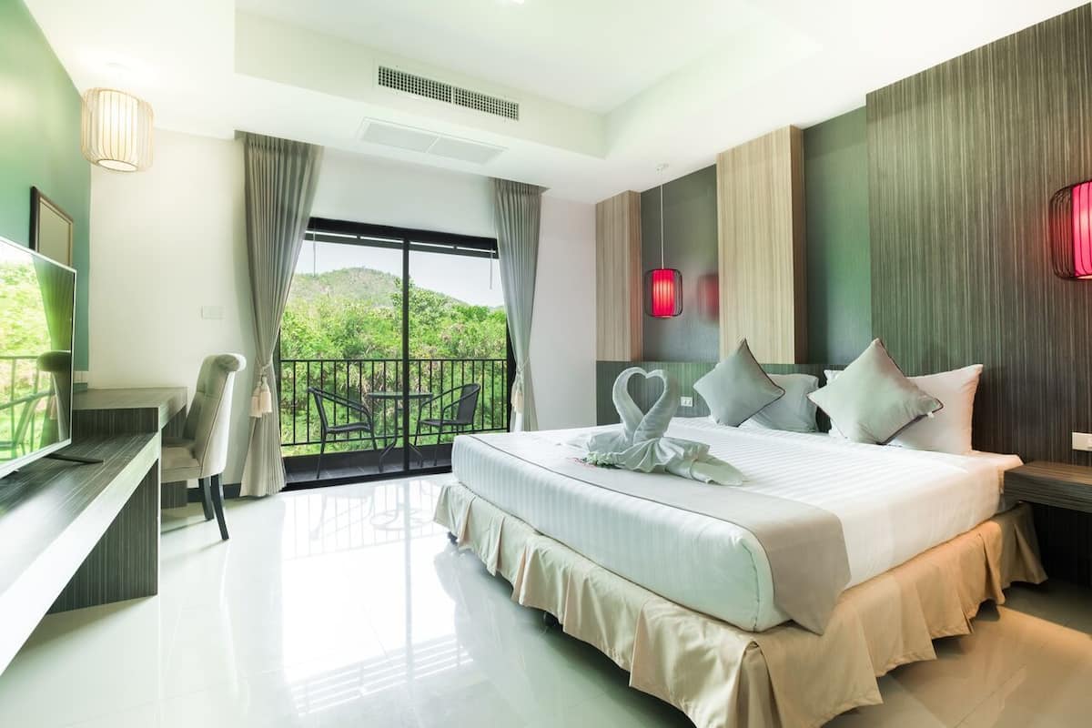 A modern hotel room with a large bed, a desk, and a balcony with a view of greenery.
