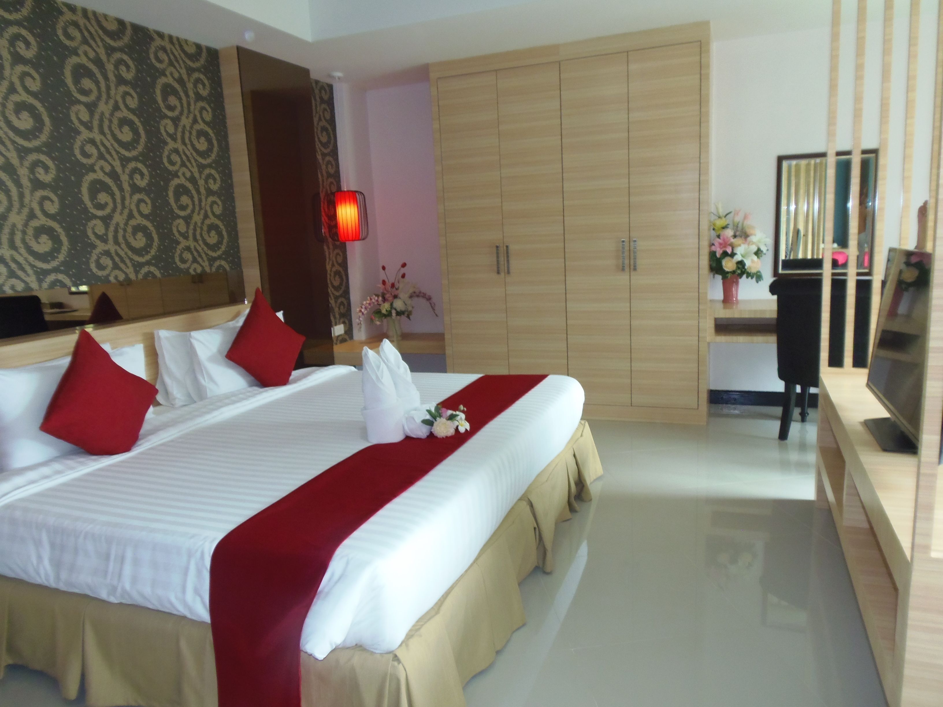A hotel room with a large bed, red pillows, a red and white bedspread, a wooden wardrobe, a desk with a computer, and a mirror.