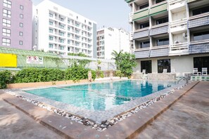 Une piscine dont le bord est recouvert de carreaux, entourée d’une bordure basse en pierre. Au fond, on aperçoit des immeubles à plusieurs étages dotés de balcons.