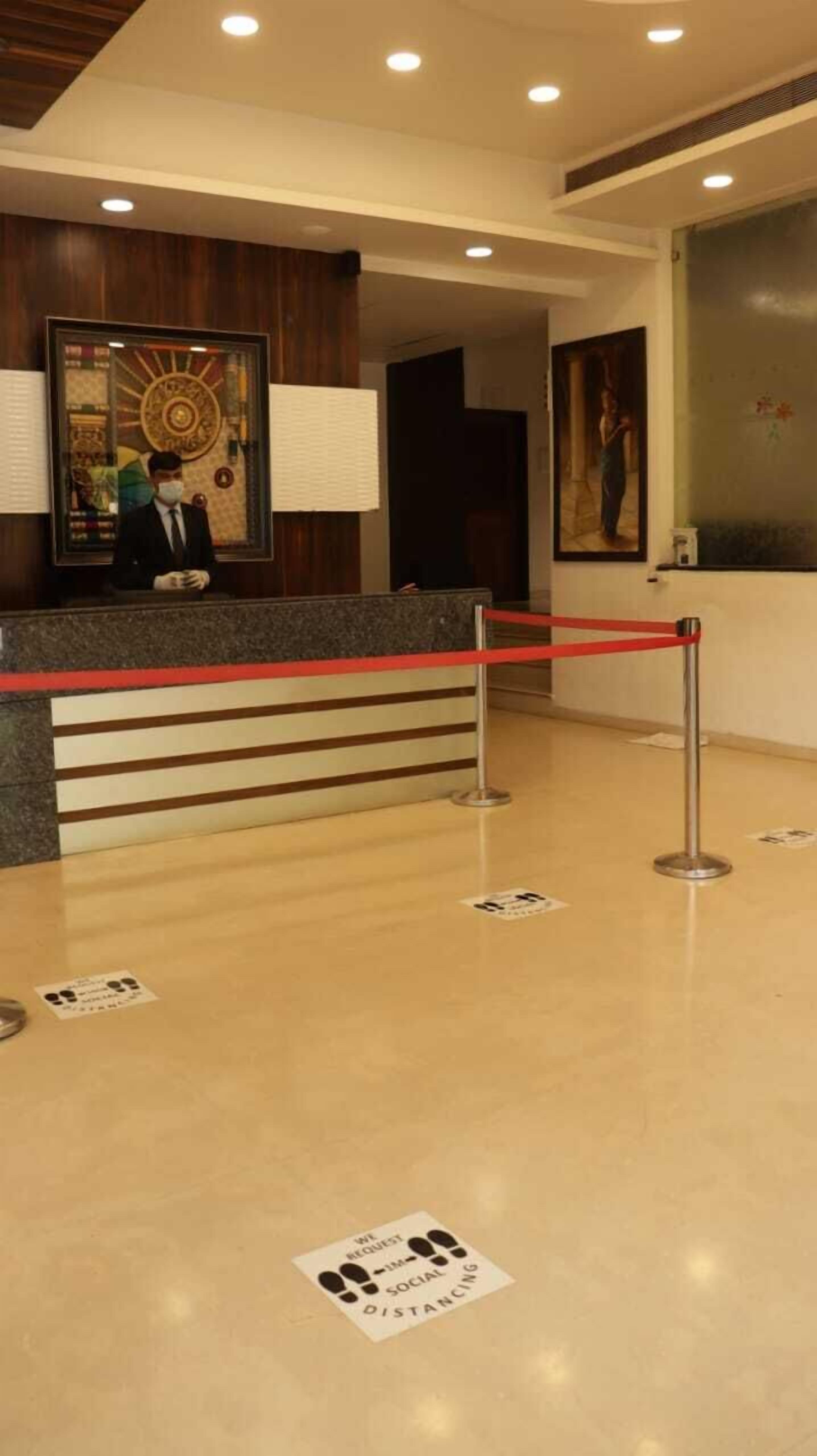 A hotel reception area with a wooden reception desk, a red rope barrier, and a floor sign requesting social distancing.
