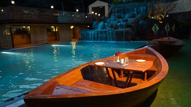 Ein hölzernes Boot mit Tisch und Stühlen an einem Pool, im Hintergrund ein Wasserfall.