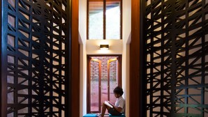 Un estrecho pasillo con puertas de celosía de madera y una persona sentada en un banco leyendo un libro.