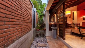 Un estrecho callejón con un muro de ladrillo a un lado y un edificio moderno al otro. En el centro, hay un pequeño jardín con plantas y una estatua.