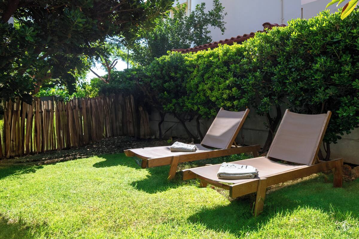 Two wooden lounge chairs with cushions and towels on a grassy lawn, surrounded by a wooden fence and lush greenery.