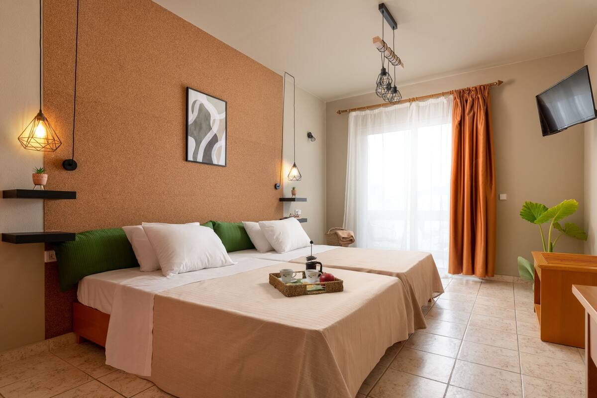 A hotel room with two beds, a flat-screen TV, a large window with curtains, and a small plant.