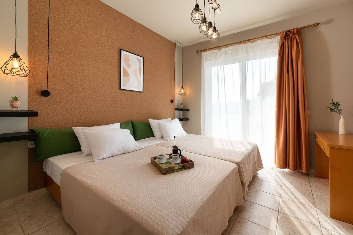 A hotel room with two beds, a breakfast tray, a green pillow, a white pillow, a framed picture, a hanging light fixture, and a window with curtains.