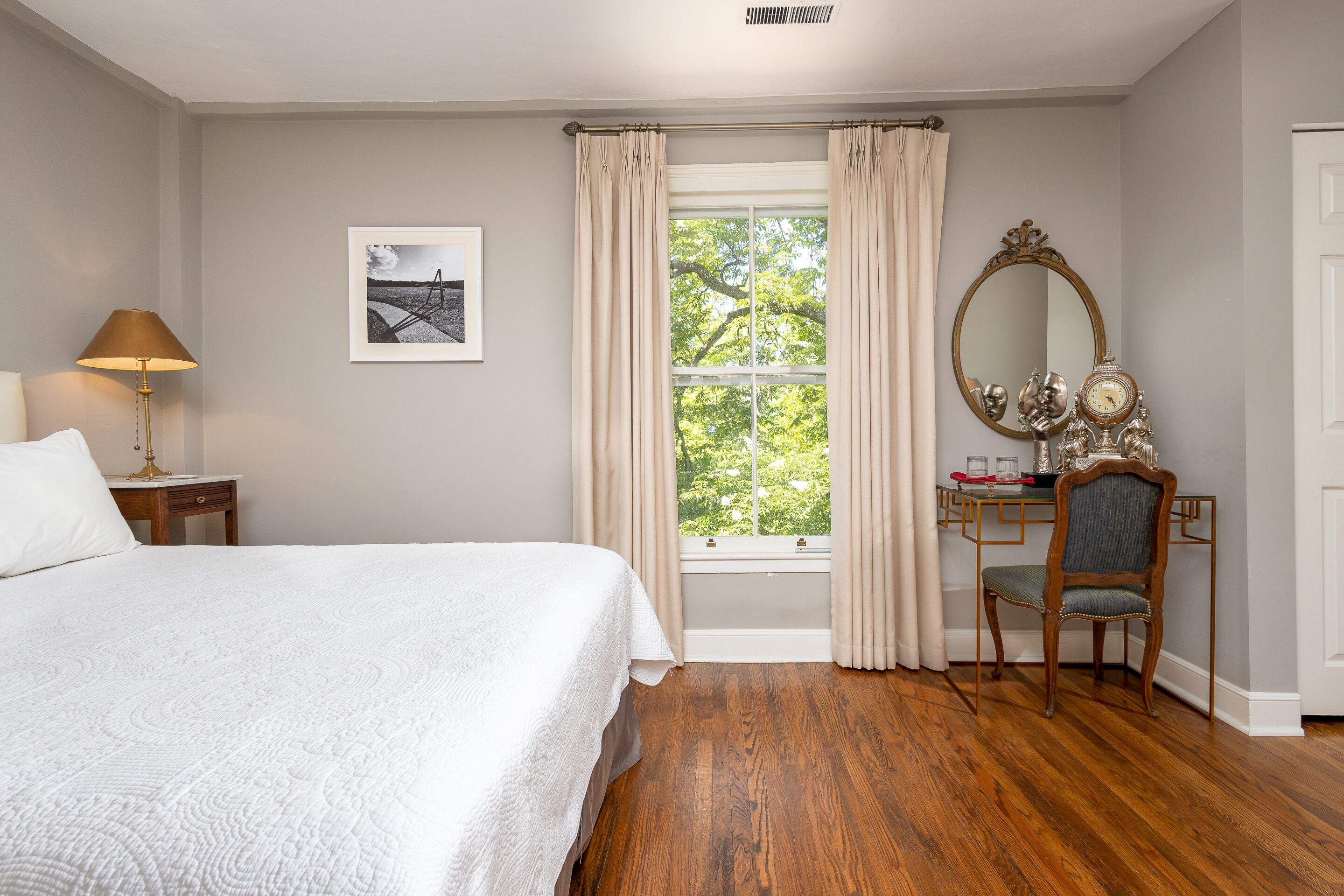 Un dormitorio con cama, escritorio, silla, espejo y ventana con cortinas.