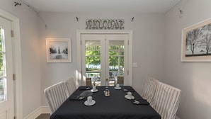 Un coin repas avec une nappe noire, des chaises à rayures, une pancarte « BIENVENUE », des photos encadrées et une vue sur l’extérieur à travers la porte.