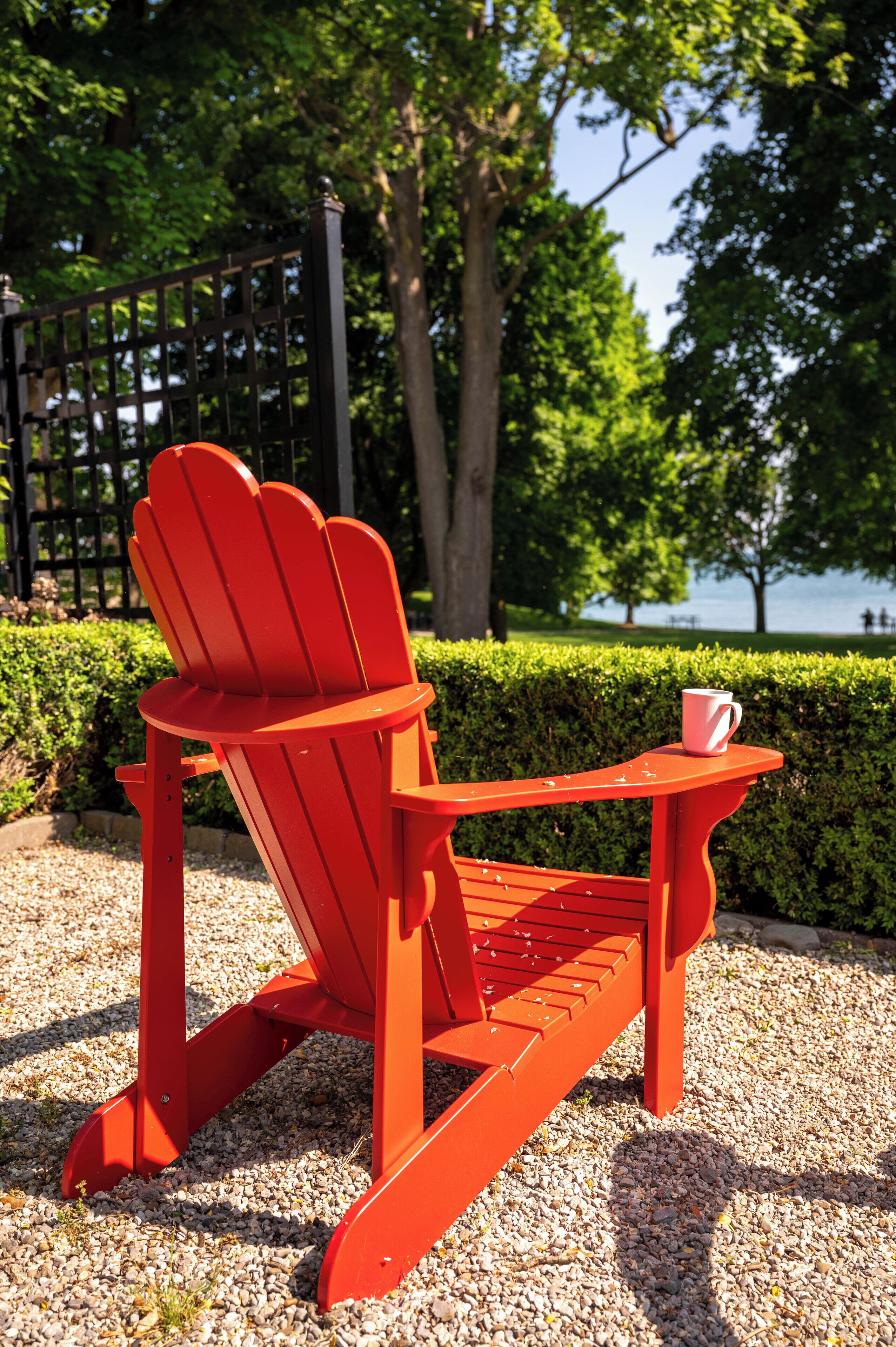 A red Adirondack chair with a white mug on a small table, set on a gravel surface with a hedge and trees in the background.