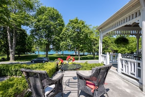 An outdoor patio with wicker furniture, a round table, and a view of a lake. - The Old Bank House Bed & Breakfast (Niagara-on-the-Lake)
