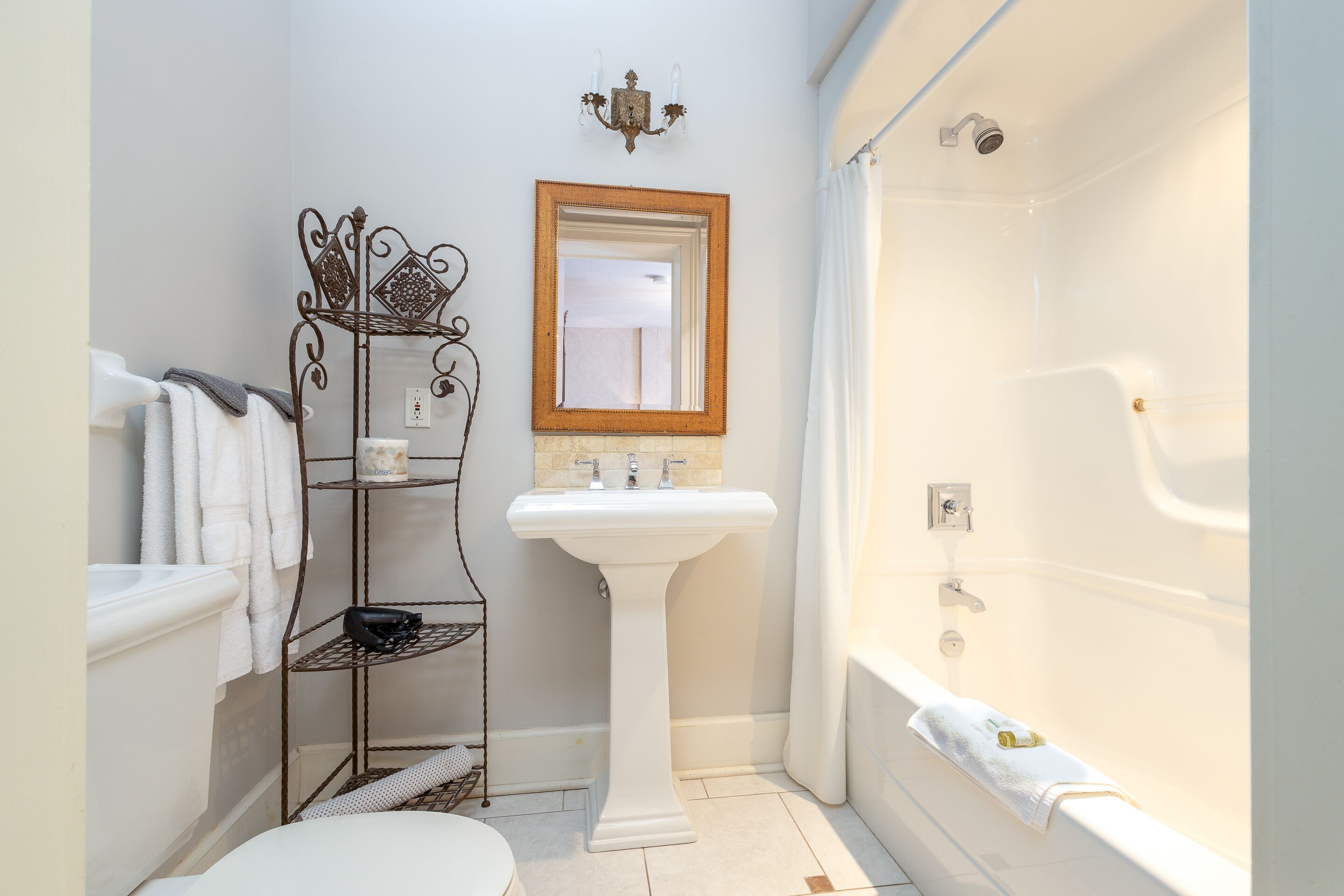A bathroom with a bathtub, a pedestal sink, a vanity with a mirror, and a metal shelf unit.