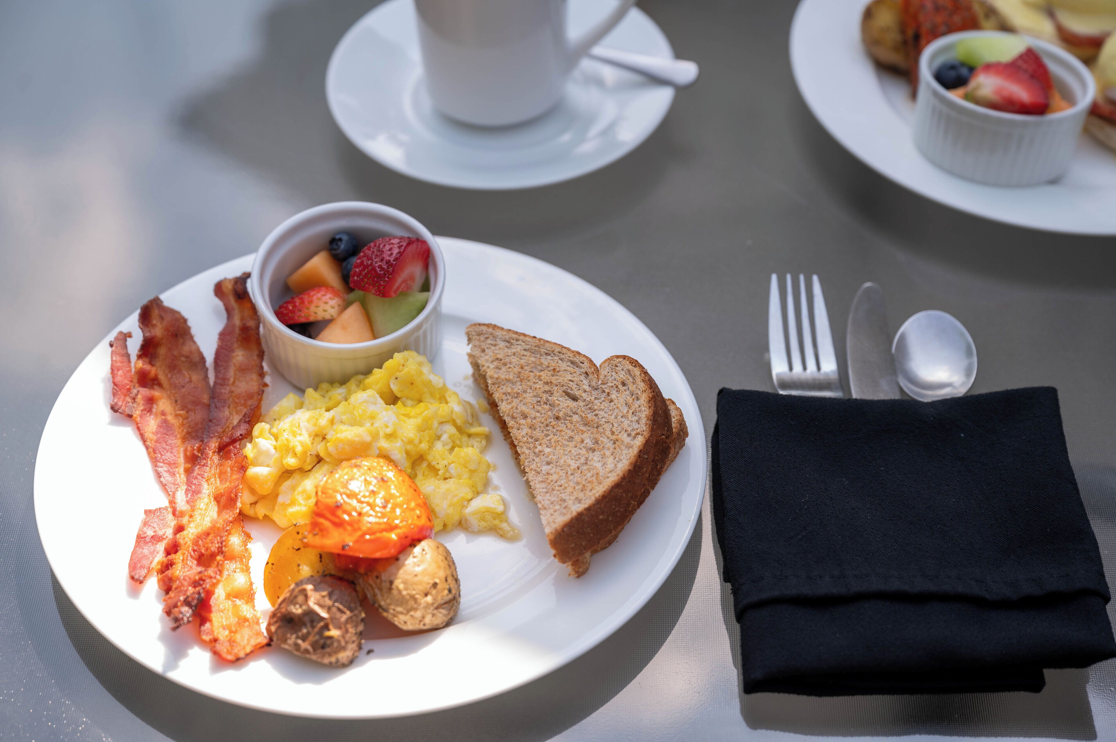 A breakfast plate with bacon, eggs, toast, and a fruit bowl.