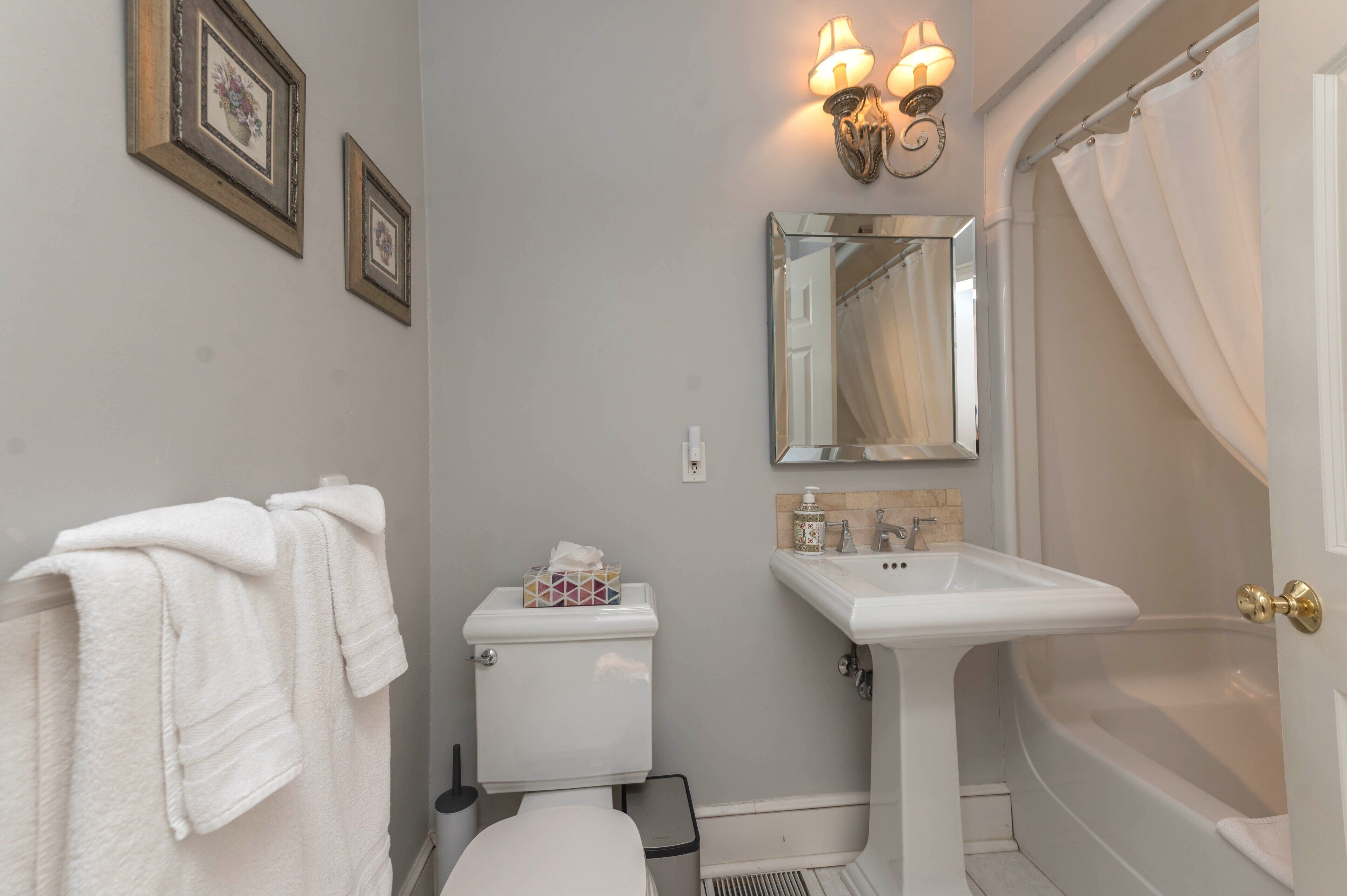 A bathroom with a toilet, sink, and bathtub. There are towels hanging on a rack, a mirror, and framed pictures on the wall.