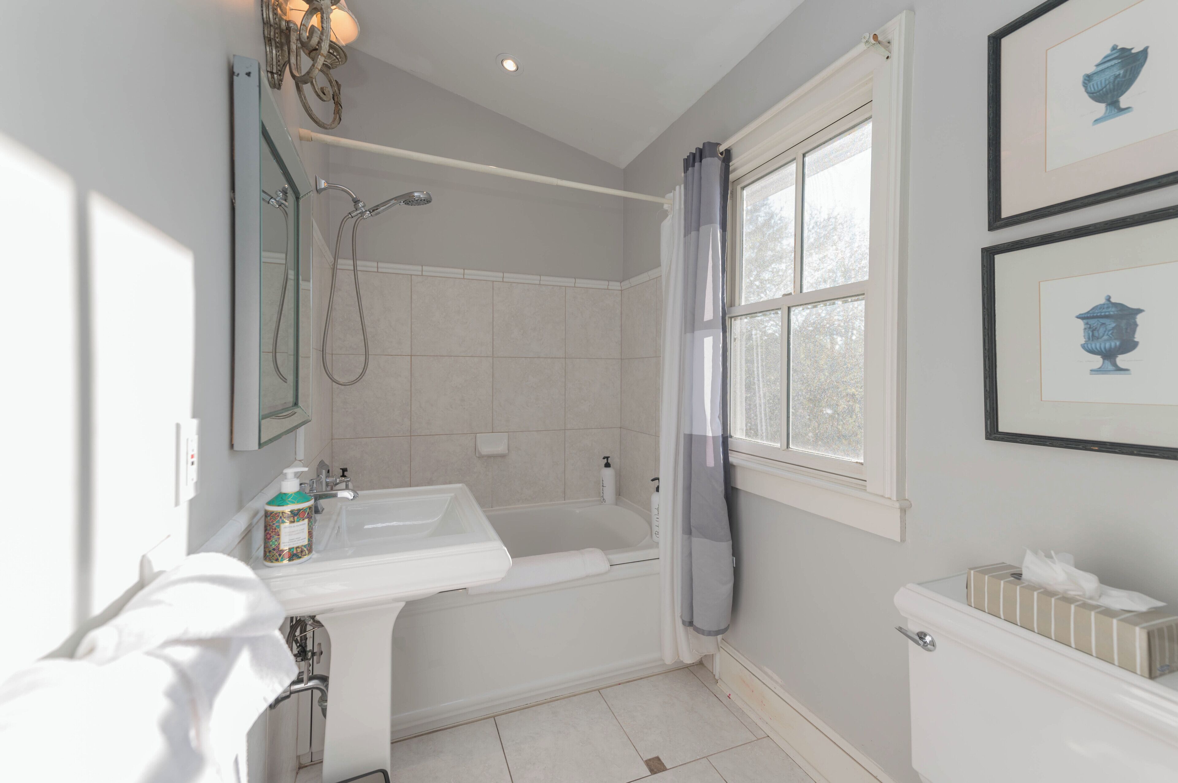 A modern bathroom with a bathtub, a sink, and a toilet. There is a window with curtains, a mirror, and framed artwork on the wall.