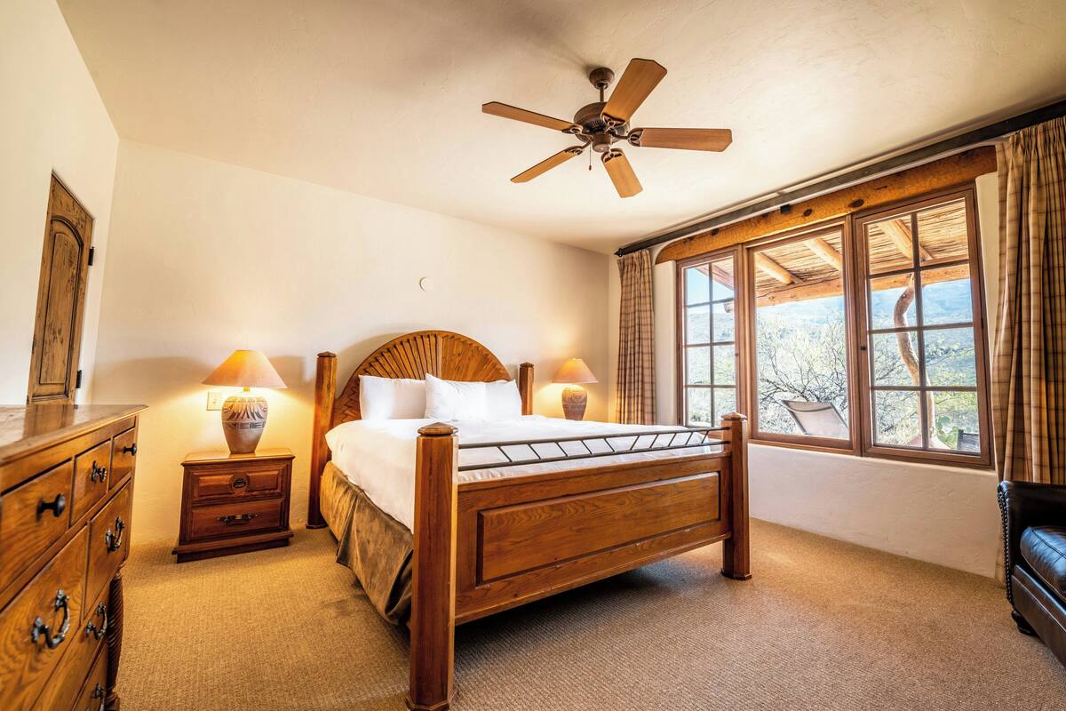 A bedroom with a wooden bed, two bedside tables with lamps, a ceiling fan, and large windows with curtains.