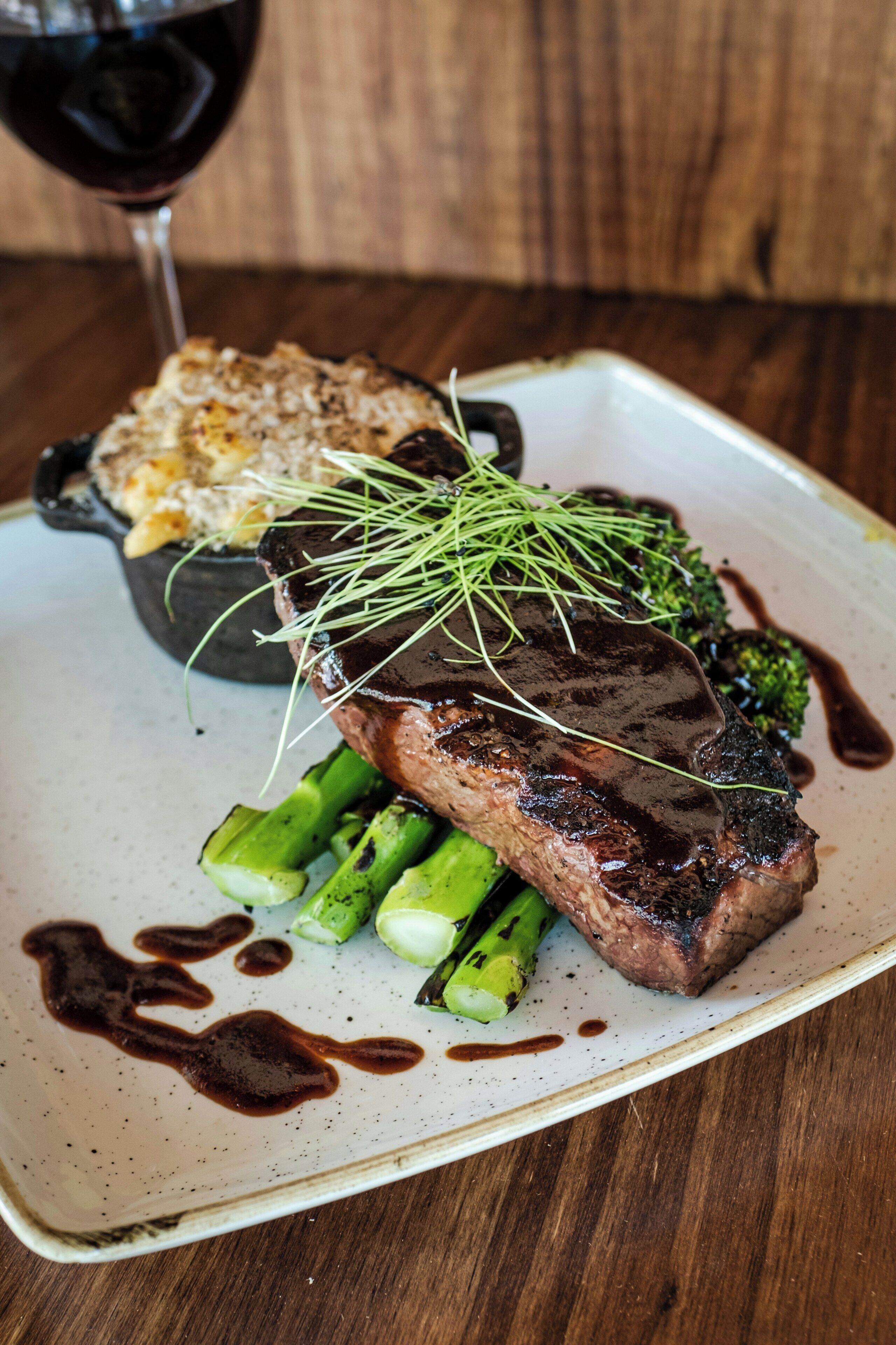 A plate with a steak, asparagus, and a side dish, with a glass of red wine in the background.