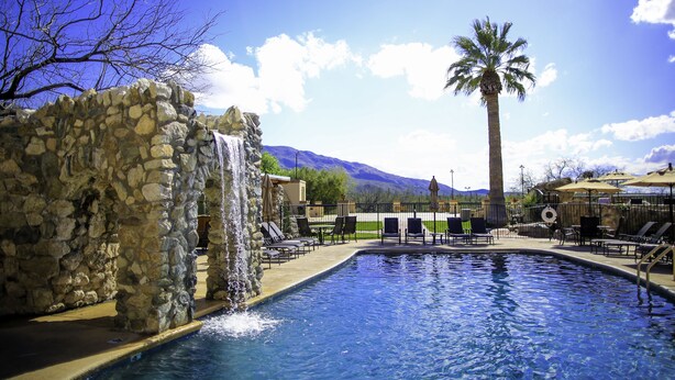 A pool with a stone wall and waterfall feature, surrounded by lounge chairs and umbrellas, with mountains in the background. - Tanque Verde Ranch (Tucson)