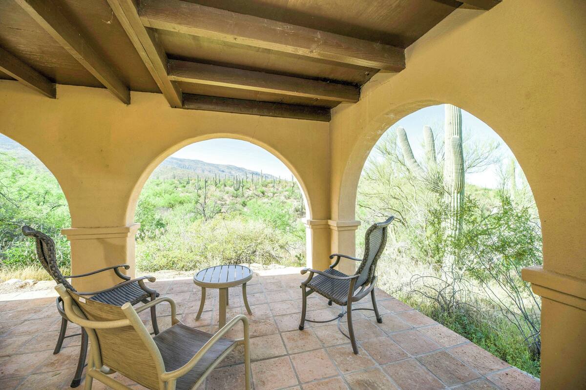 An outdoor patio with two chairs and a table, surrounded by arches with a view of desert vegetation.