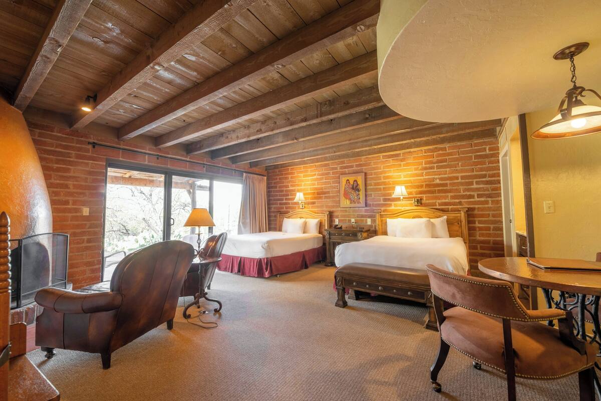 A hotel room with a brick wall, wooden ceiling beams, two beds, a desk, a chair, and a table.