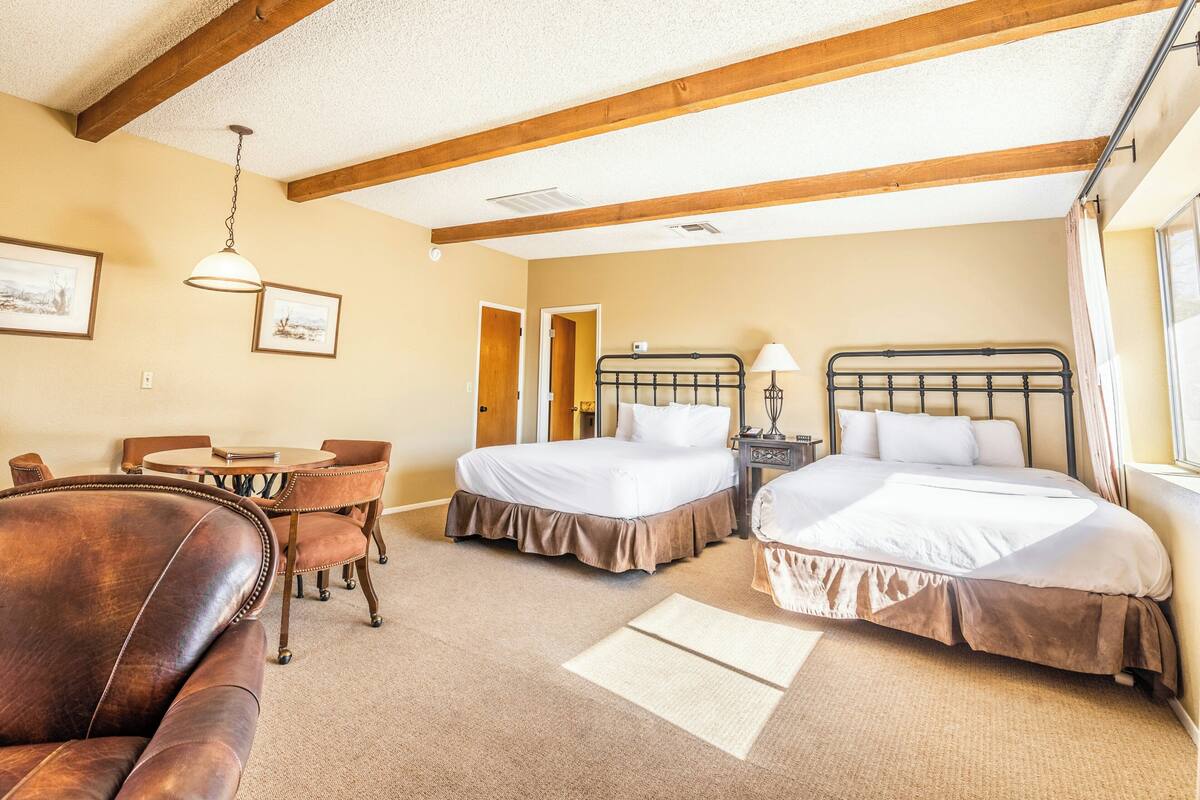 A hotel room with two beds, a desk, chairs, and a hanging light fixture.